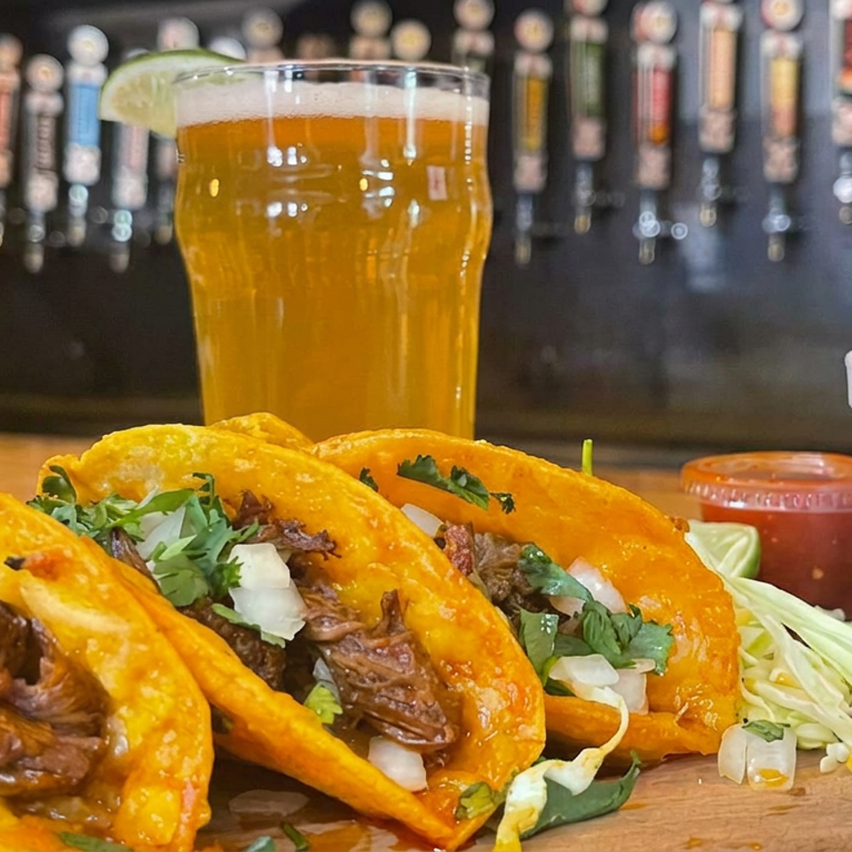 Three birria tacos topped with cilantro and onions sit beside a lime-garnished beer, cabbage slaw, salsa, and a cup on a bar.