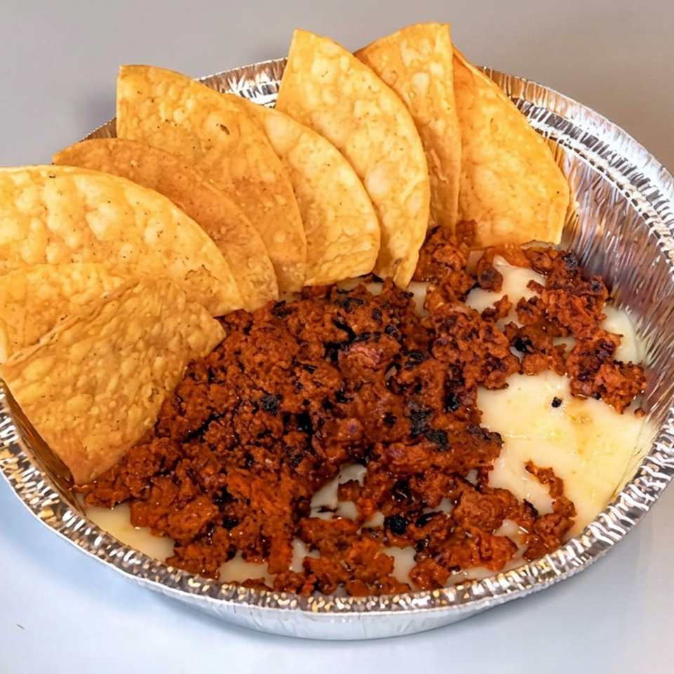 Foil tray filled with creamy cheese dip, seasoned ground meat, and golden tortilla chips arranged around the edge.